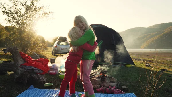 Two little blonde female children sisters hugging near campfire and tent