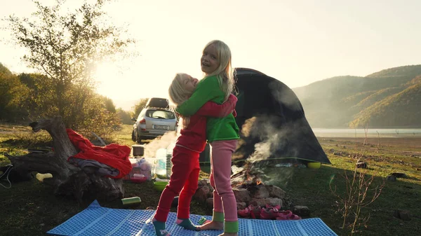 Two little blonde female children sisters hugging near campfire and tent