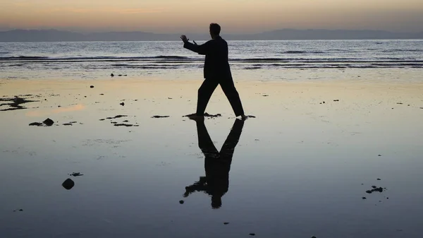 Gün batımında deniz kenarında qigong egzersizi yapan bir adamın silueti.