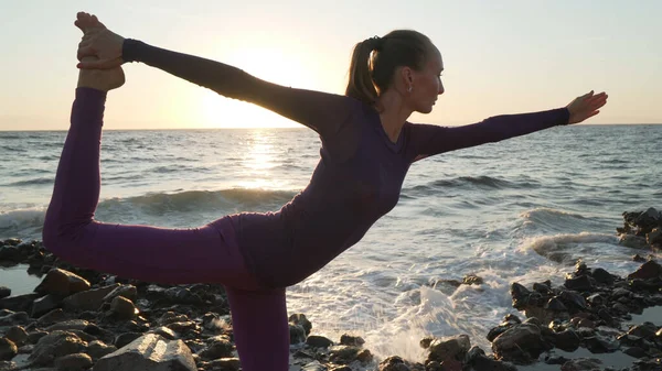 Joven hembra realiza un arco de pie tirando de yoga pose en la orilla del mar . —  Fotos de Stock