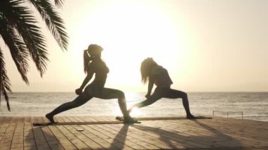 İki kadının silueti sahilde gün ışığında savaşçı yoga pozisyonu alıyor.