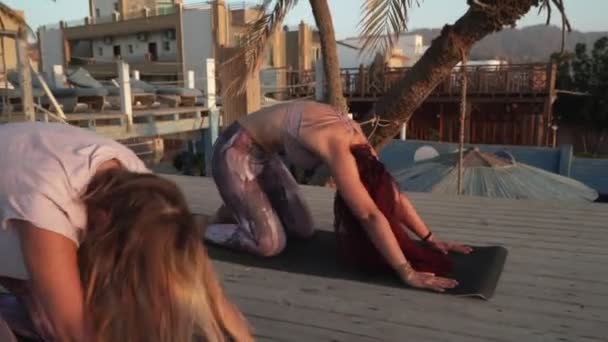 Dos mujeres practican yoga en la zona del salón del hotel junto al mar al atardecer — Vídeos de Stock