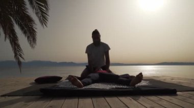 Güneş doğarken deniz kenarında bir kadın için Tayland yogası masajı yapan masör silueti.
