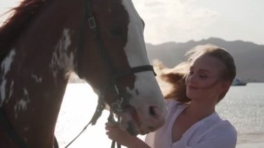 Güzel beyaz kız gün doğumunda ağır çekimde deniz kıyısında bir atı okşuyor.