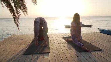 İki kadın gün batımında deniz kıyısındaki tatil köyünde farklı yoga asanas alıştırmaları yapıyor.