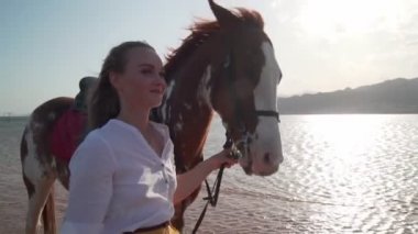 Kafkasyalı genç bir kız gün batımında sahilde ağır çekimde yürüyen bir atı okşuyor.