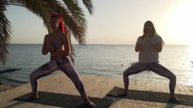 İki kadının silueti, gün ışığında deniz kenarında Tanrıça yogası pozisyonu alıyor.