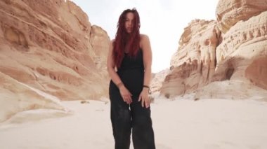 Siyah takım elbiseli genç kızıl kız kumlu çölde coşkulu dans ediyor.