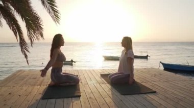 İki kadının silueti gün batımında deniz kenarında meditasyon yapıyor.