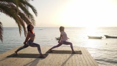 İki kadın gün batımında deniz kenarındaki iskelede yoga yapıyor.