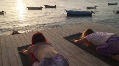 İki kadın gün batımında deniz kenarında yoga kedi duruşu yapıyor.