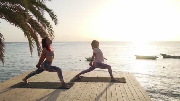 Две женщины практикуют йогу воина на приморском пирсе на закате — стоковое видео