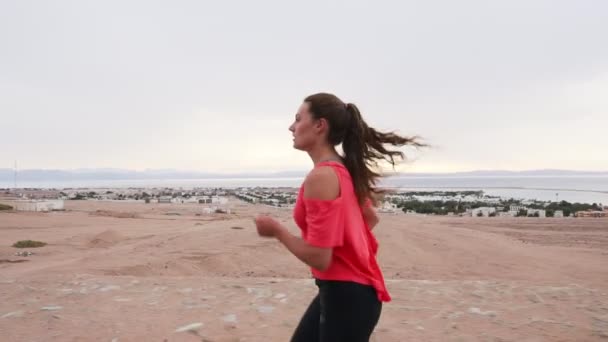 Vue de côté de brune caucasienne fille jogging le long de la route du désert au ralenti — Video