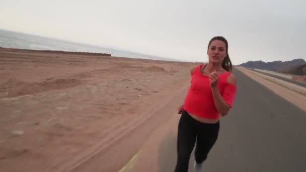 Chica joven corriendo a lo largo de la carretera en la orilla del mar dinámico movimiento de la cámara activa — Vídeo de stock