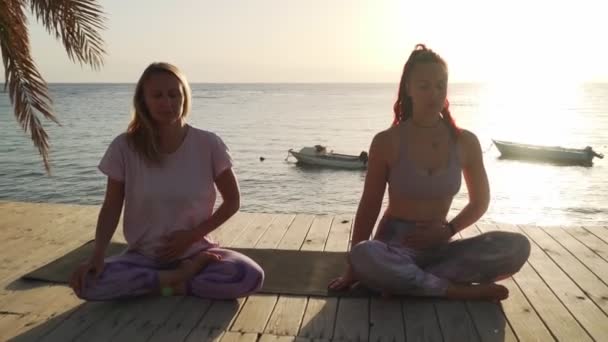 Две кавказские девушки делают дыхательные упражнения на берегу моря под солнцем — стоковое видео