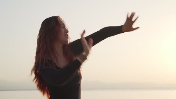 Pelirroja en traje negro practica danza extática en la costa del mar al amanecer — Vídeos de Stock