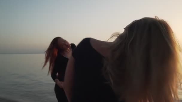 Siyah takım elbiseli iki kız, gün ışığında deniz kıyısında coşkulu bir şekilde dans ediyorlar. — Stok video