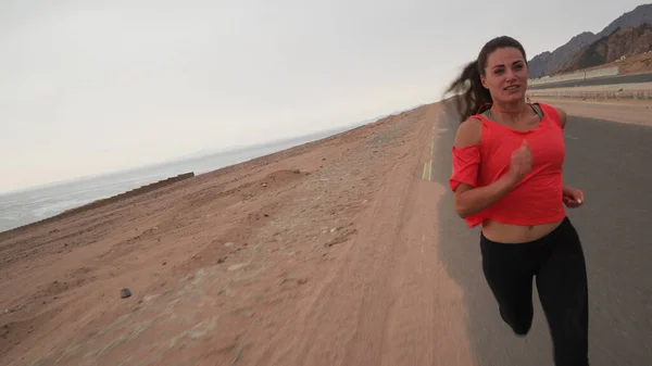 Esmer, beyaz bir kadının deniz kıyısındaki yolda koşarkenki açılı görüntüsü.