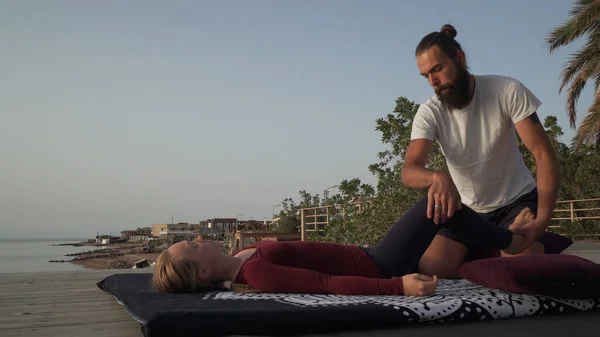 Güneş batarken deniz kıyısında bir kadın için Tayland yogası yapan yakışıklı sakallı adam..