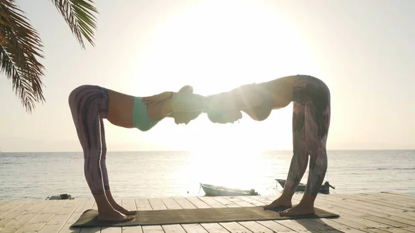 İki kadın, gün ışığında deniz kenarında omuz açıcı yoga egzersizi yapıyor.