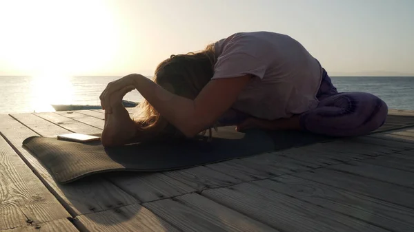 Mujer flexible practica mahamudra yoga a orillas del mar bajo el sol —  Fotos de Stock