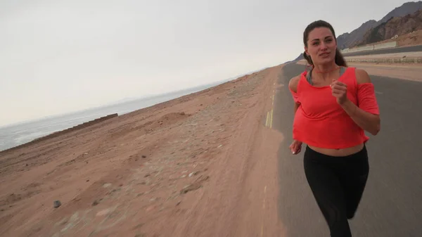 Esmer, beyaz bir kadının deniz kıyısındaki yolda koşarkenki açılı görüntüsü.