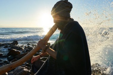 View of man sitting on seashore in sunlight and playing didjireedoo instrument. clipart