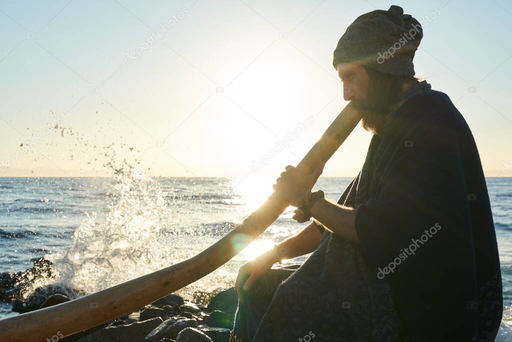 Bearded man playing his didgeridoo Australian instrument in sunshine at seashore.