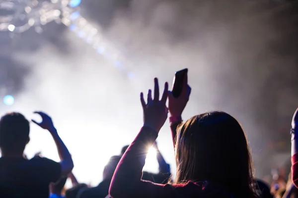 Audience with hands raised at a music festival and lights streaming down from above the stage. Soft focus, high ISO, grainy image. — Stock Photo, Image