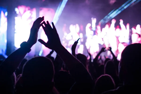 Audience with hands raised at a music festival and lights streaming down from above the stage. Soft focus, high ISO, grainy image. — Stock Photo, Image