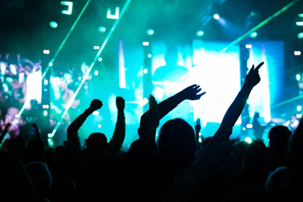 Audience with hands raised at a music festival and lights streaming down from above the stage. Soft focus, high ISO, grainy image. — Stock Photo, Image