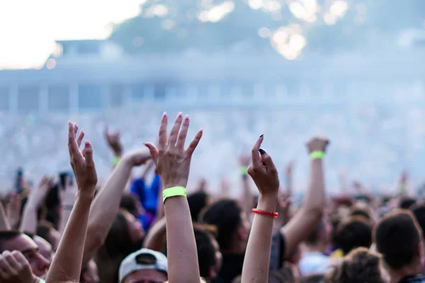Audience with hands raised at a music festival and lights streaming down from above the stage. Soft focus, high ISO, grainy image. — Stock Photo, Image