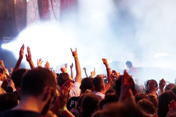 Audience with hands raised at a music festival and lights streaming down from above the stage. Soft focus, high ISO, grainy image. — Stock Photo, Image