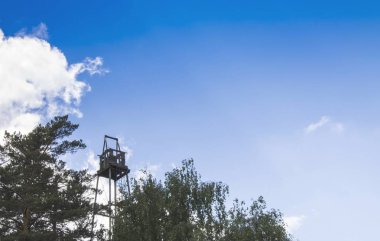 Mavi gökyüzü ve beyaz bulutlara karşı yeşil bir ormanın üzerinde eski yangın gözetleme kulesi