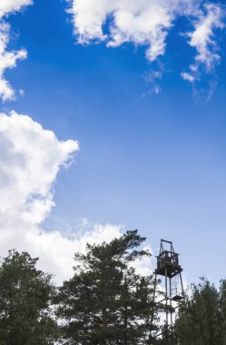 Mavi gökyüzü ve beyaz bulutlara karşı yeşil bir ormanın üzerinde eski yangın gözetleme kulesi