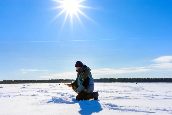 Winter fishing. Ice fisherman fishing in the winter on the river. winter fisherman sits in camouflage clothing near a hole with an ice drill on a frozen snow covered river on a winter day. — Stock Photo, Image