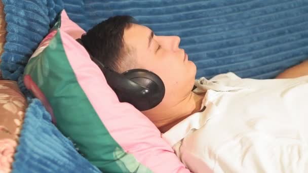 若い男がリラックスした音楽、穏やかな音楽を聞いて自宅でヘッドフォンを閉じてソファの上に休んでいる。リラックス心の平安ヘッドフォンと音楽を聴き — ストック動画