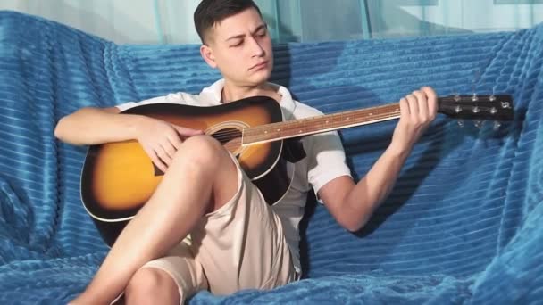 Junger Kerl sitzt auf der Couch und spielt Gitarre. vor die Kamera. Gitarre spielen lernen — Stockvideo