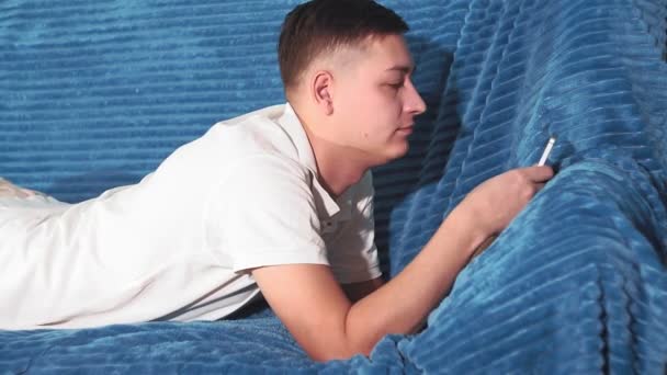 若い男がソファに横になって電話を見てる電子メール、インターネット、ソーシャルネットワークのチェック — ストック動画