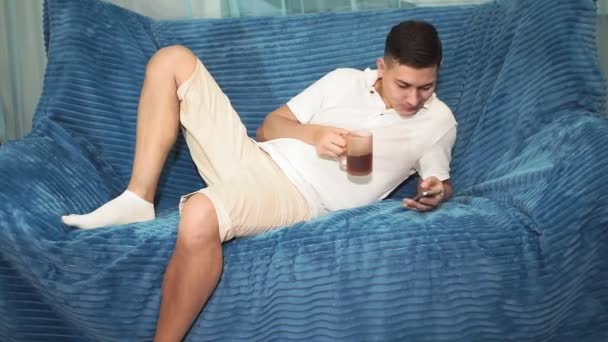 Jovem está deitado no sofá e olha para o telefone, bebendo chá. Verifica e-mail Internet, redes sociais — Vídeo de Stock