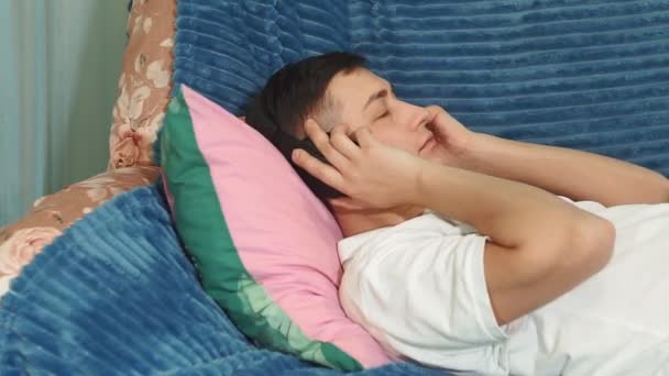 Joven hombre milenario con auriculares en la cabeza tumbado en el sofá escucha música tranquila y relajante — Vídeo de stock