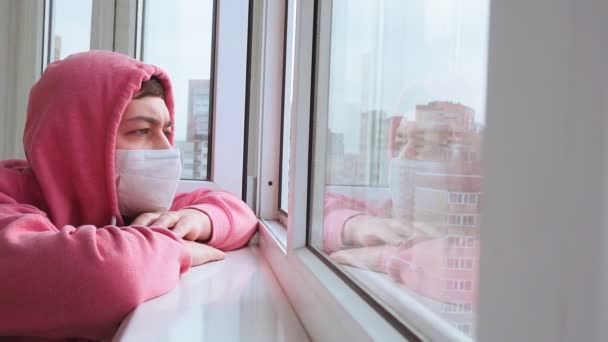 Un hombre con una máscara protectora en casa. máscara médica estéril mira por la ventana y está en cuarentena en casa y auto-aislamiento durante la epidemia de coronavirus — Vídeo de stock