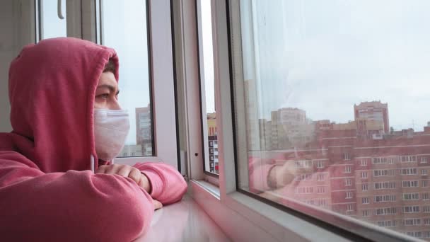 家の保護マスクの男。隔離隔離中の窓には顔をコロナウイルス感染から守るために外科用の医療用マスクをつけています — ストック動画