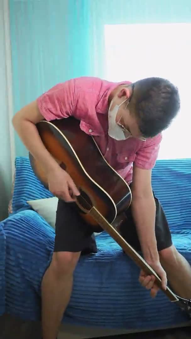 Mladý muž hraje na akustickou kytaru v masce před virem. Svislé video — Stock video
