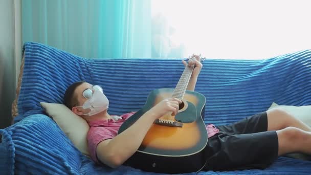 Молодой человек играет на гитаре в маске от коронавирусной инфекции и очков . — стоковое видео