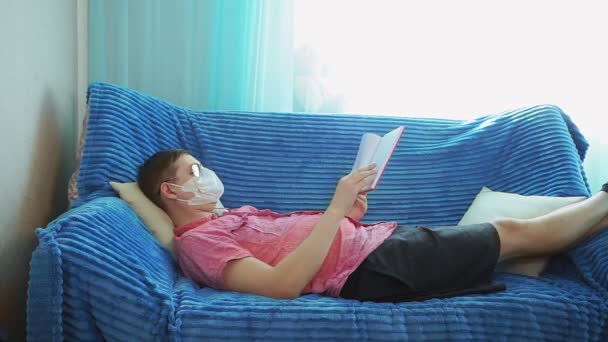 Junger Mann liest zu Hause ein Buch in einer Maske vor dem Coronavirus, Selbstisolierung — Stockvideo