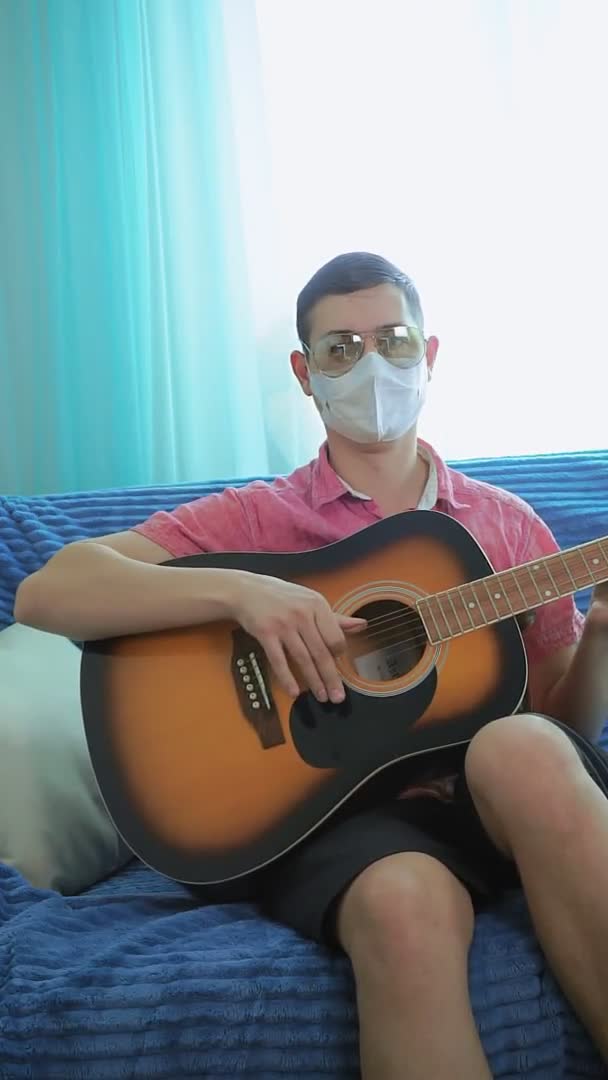 젊은 남자가 안경을 끼고 바이러스 마스크를 쓰고 음향 기타를 연주 합니다. 수직 영상 — 비디오