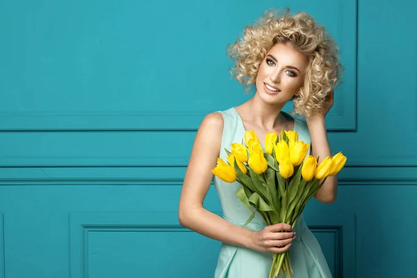 Vacker blond flicka i blå klänning med blommor tulpaner i händerna på en turkos bakgrund — Stockfoto