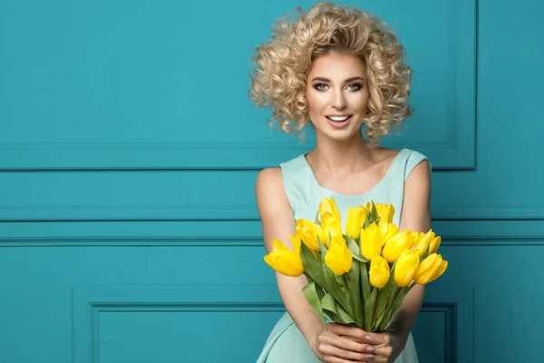 Mooi blonde meisje in de blauwe jurk met bloemen tulpen in handen op een turkooizen achtergrond — Stockfoto