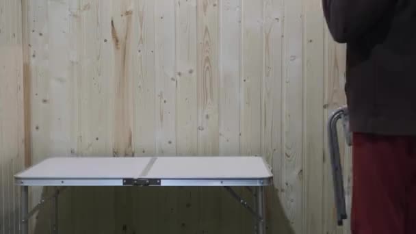 The guy put a folding stool in front of the table and sits on it — Stock Video